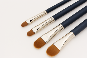 Four premium makeup brushes in graduated sizes displayed diagonally on a clean white background, featuring navy blue handles, silver ferrules, and soft brown synthetic bristles.