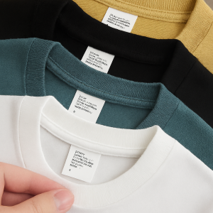 Stack of four crewneck t-shirts in teal, mustard yellow, black, and white, laid on a wooden surface with a hand holding the white shirt’s collar.