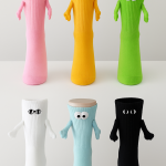 Six novelty socks in pink, yellow, green, white, light blue, and black, each standing upright with googly or embroidered eyes and matching fabric arms, arranged in a 2x3 grid on a white background.