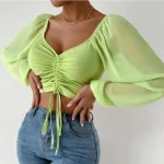 Woman wearing a green ruched crop top with sheer long sleeves, square neckline, and tie-front detail, styled with high-waisted jeans and a white clutch.