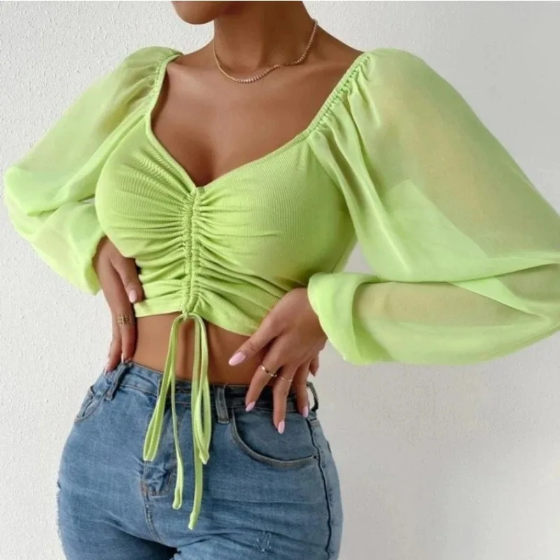 Woman wearing a green ruched crop top with sheer long sleeves, square neckline, and tie-front detail, styled with high-waisted jeans and a white clutch.