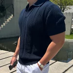 Man wearing a navy blue ribbed mesh polo shirt with white pants, standing near a modern concrete structure and water feature