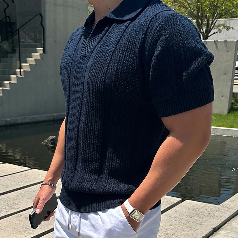 Man wearing a navy blue ribbed mesh polo shirt with white pants, standing near a modern concrete structure and water feature