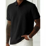 Model wearing a black chevron-textured polo shirt with short sleeves, white pants, tattoos on both arms, and a silver watch, standing indoors against a white wall.