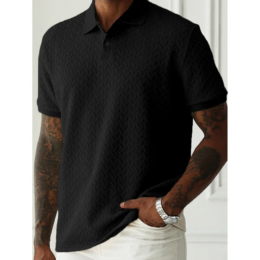 Model wearing a black chevron-textured polo shirt with short sleeves, white pants, tattoos on both arms, and a silver watch, standing indoors against a white wall.