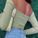 A person wearing a light green off-shoulder top and blue jeans, with a beige handbag over the shoulder, standing against a green leafy background.