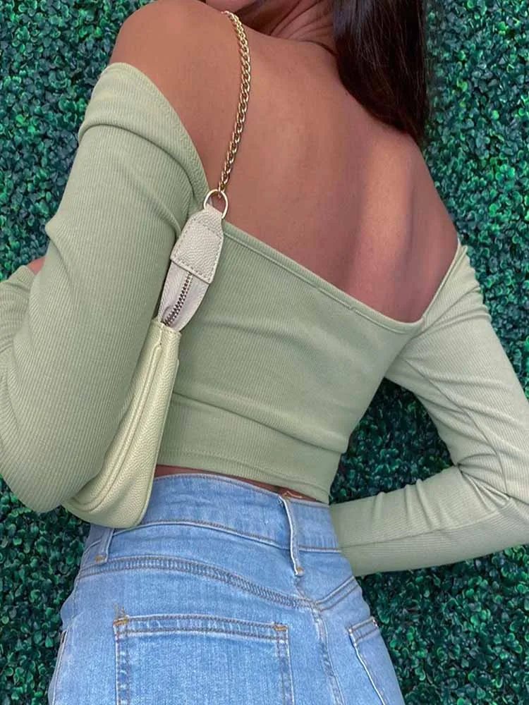 A person wearing a light green off-shoulder top and blue jeans, with a beige handbag over the shoulder, standing against a green leafy background.
