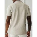 Back view of a model wearing a beige chevron-textured polo shirt with short sleeves, tattoos on both arms, and a silver wristwatch, paired with white pants.