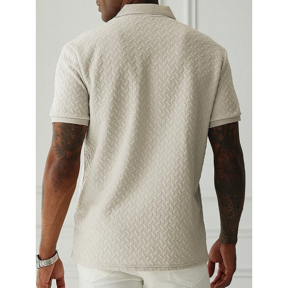 Back view of a model wearing a beige chevron-textured polo shirt with short sleeves, tattoos on both arms, and a silver wristwatch, paired with white pants.
