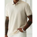 Model wearing a beige chevron-textured polo shirt with short sleeves, tattoos on both arms, and a silver watch, standing indoors with one hand in pocket.
