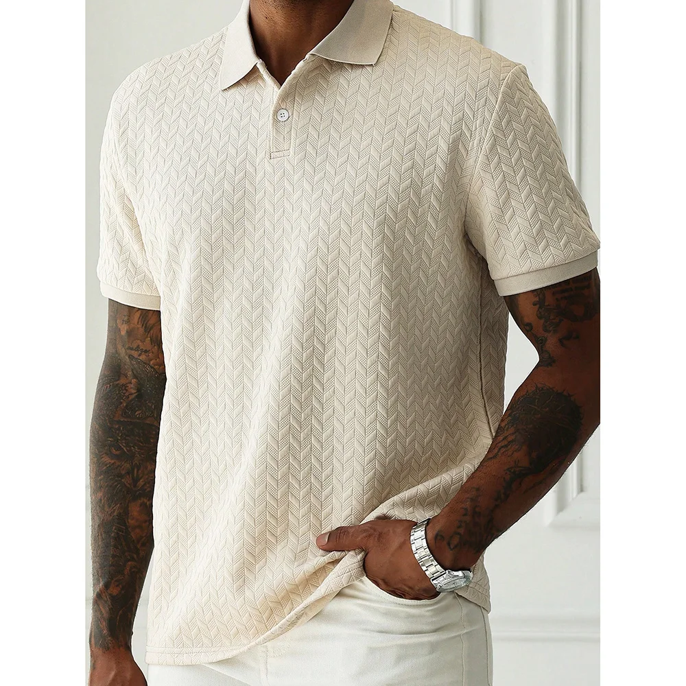 Model wearing a beige chevron-textured polo shirt with short sleeves, tattoos on both arms, and a silver watch, standing indoors with one hand in pocket.
