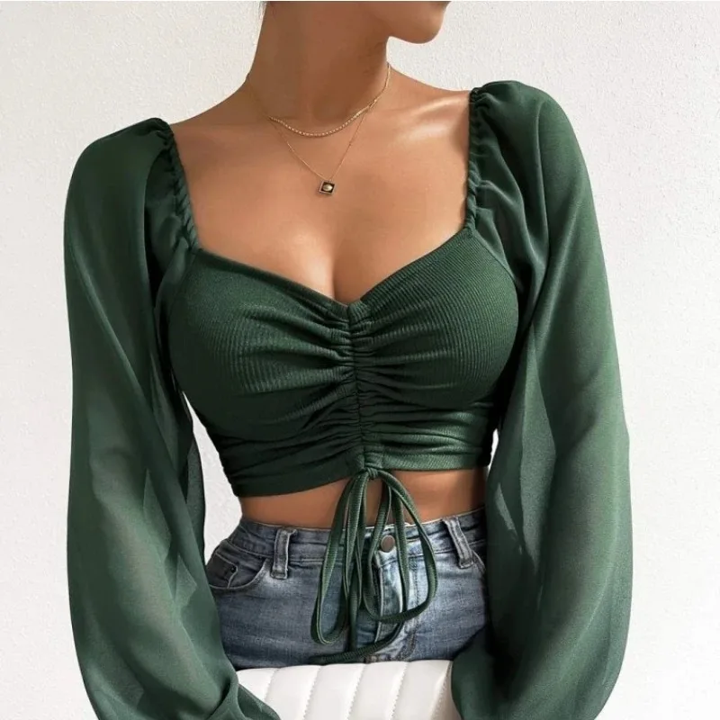Woman wearing a dark green ruched crop top with sheer long sleeves, square neckline, and tie-front detail, styled with high-waisted jeans and a gold pendant necklace.