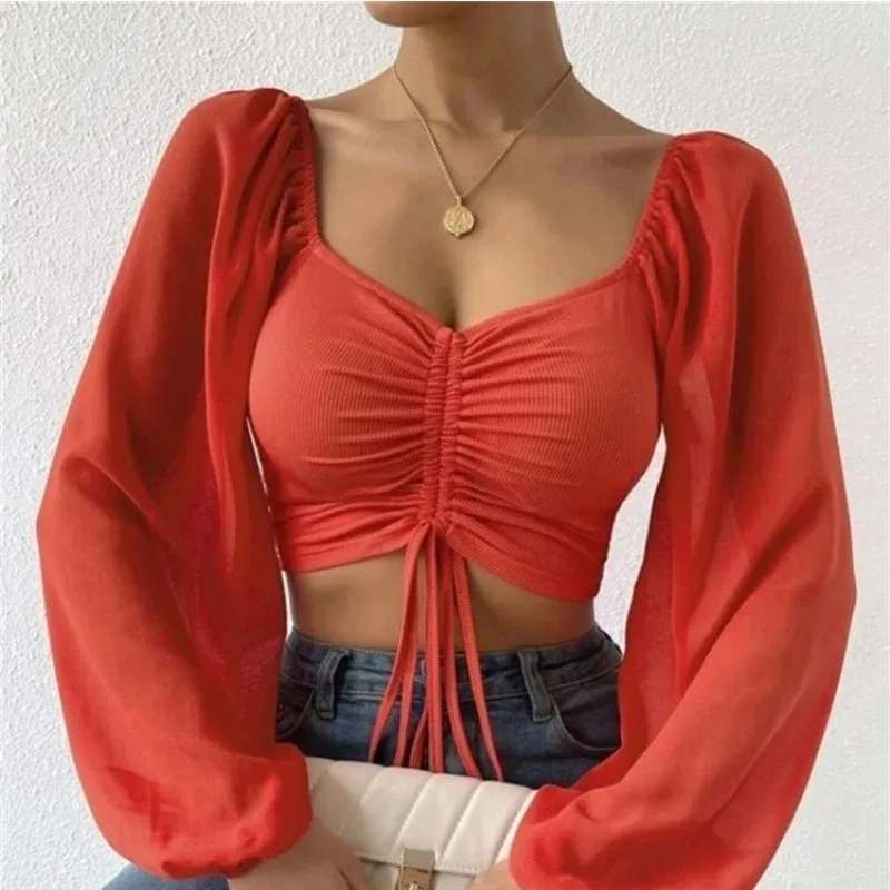 Woman wearing a red ruched crop top with sheer long sleeves, square neckline, and center drawstring detail, styled with high-waisted jeans and a white quilted clutch.