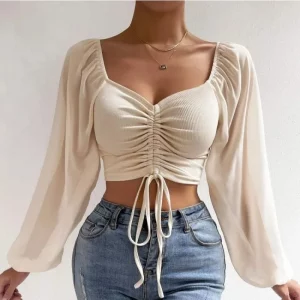 Woman wearing a beige ruched long sleeve crop top with square neckline and drawstring detail, paired with high-waisted jeans.