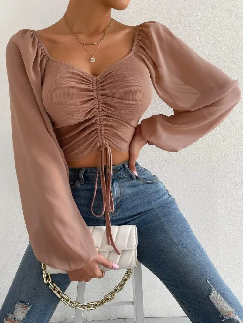 Woman wearing a light brown ruched crop top with sheer long sleeves, square neckline, and tie-front detail, styled with distressed high-waisted jeans and a white quilted handbag.