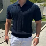 Man wearing a navy blue textured knit polo shirt with white pants, standing outdoors near concrete architecture and a water feature