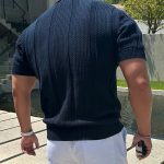 Man wearing a dark blue textured knit polo shirt and white shorts, standing outdoors near a pond and modern architecture