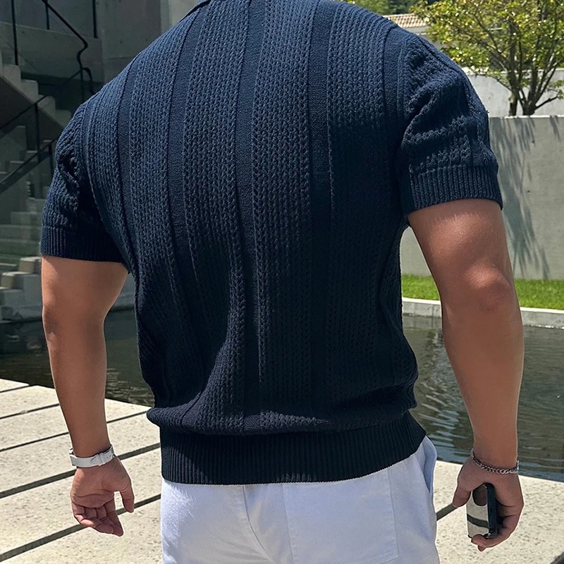 Man wearing a dark blue textured knit polo shirt and white shorts, standing outdoors near a pond and modern architecture